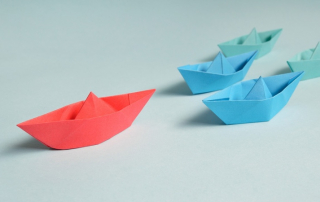 Representación del concepto de liderazgo con barcos de papel, un rojo delante y tres azules le siguen.