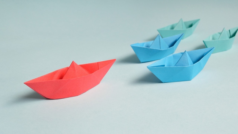 Representación del concepto de liderazgo con barcos de papel, un rojo delante y tres azules le siguen.